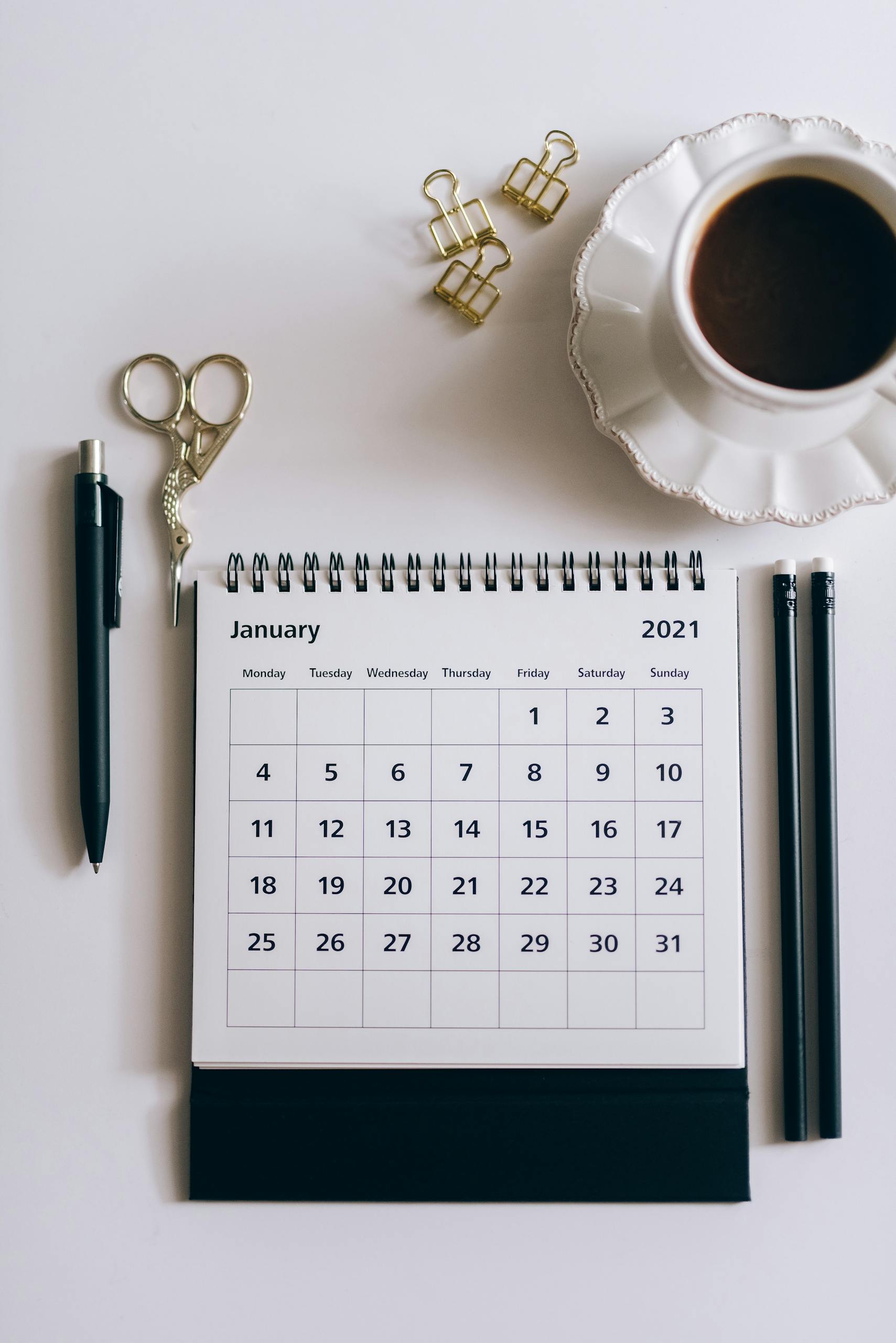 Flat lay of a 2021 desk calendar, coffee cup, and black stationery items on a white surface.